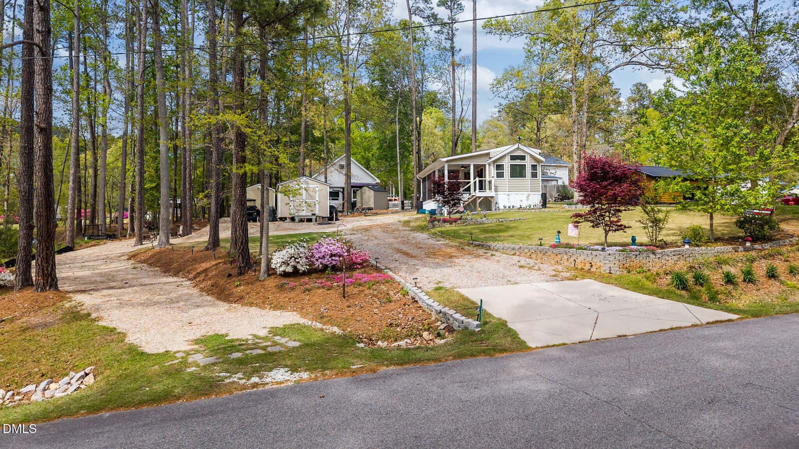 A gravel driveway and a concrete driveway lead to two single-story houses surrounded by tall trees, landscaped lawns, and flowering shrubs under a partly cloudy sky.