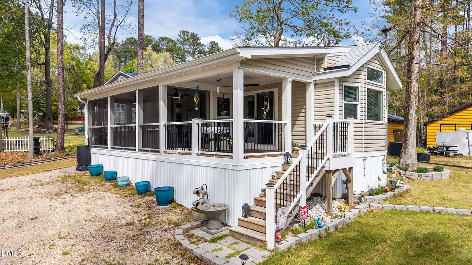 A beige manufactured home with a large screened porch, white railings, and stairs leading up to the entrance. Blue planters, a small fountain, and a gravel driveway are visible in front. Trees and neighboring houses are in the background.