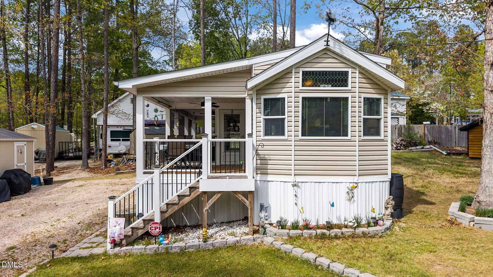 A beige manufactured home with a covered porch, steps leading up, white railing, and skirting. The yard has landscaping, small decorations, and woods in the background. A shed and a neighboring house are visible.