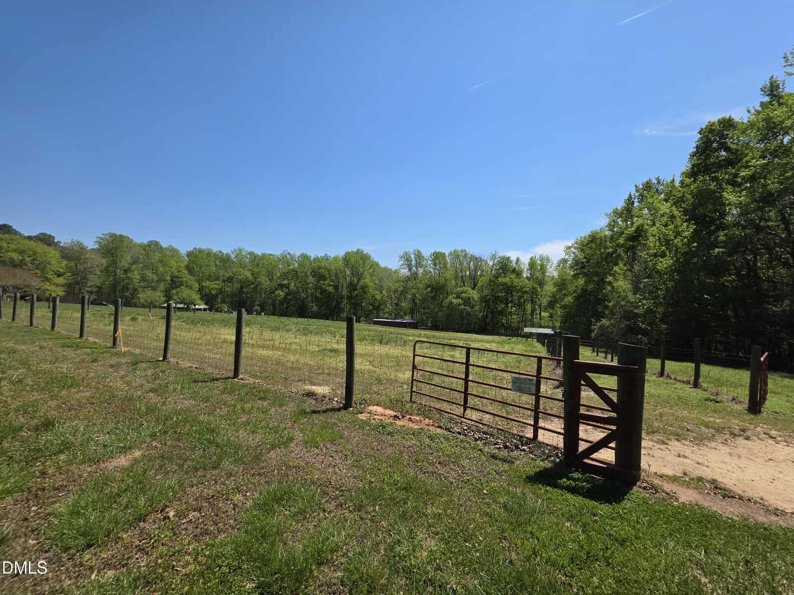 A fenced pasture on 000 Johnson Town Road with a metal gate open to a grassy field, surrounded by trees under a clear blue Zebulon sky on a sunny day. A small shed is visible in the distance.