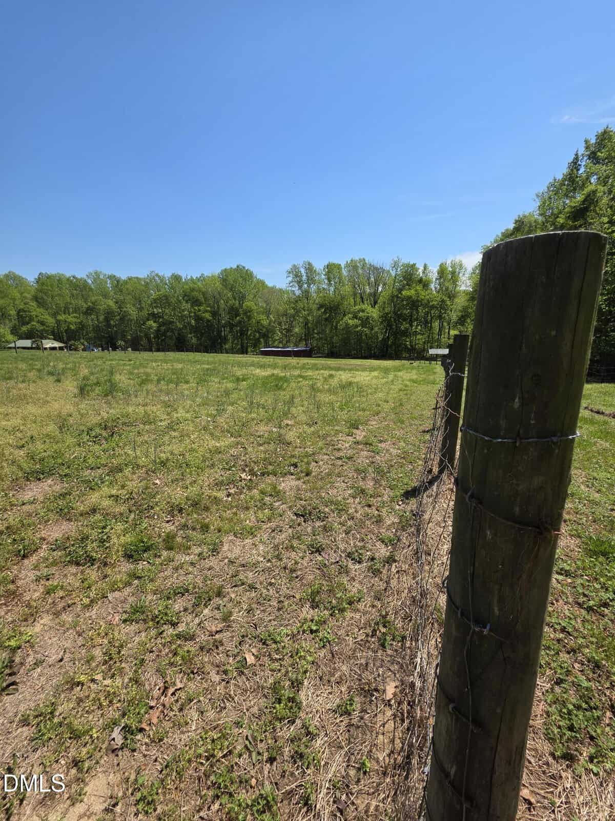 A wooden fence post with wire fencing stands in the foreground of a grassy field under a clear blue sky, bordered by green trees in the background.