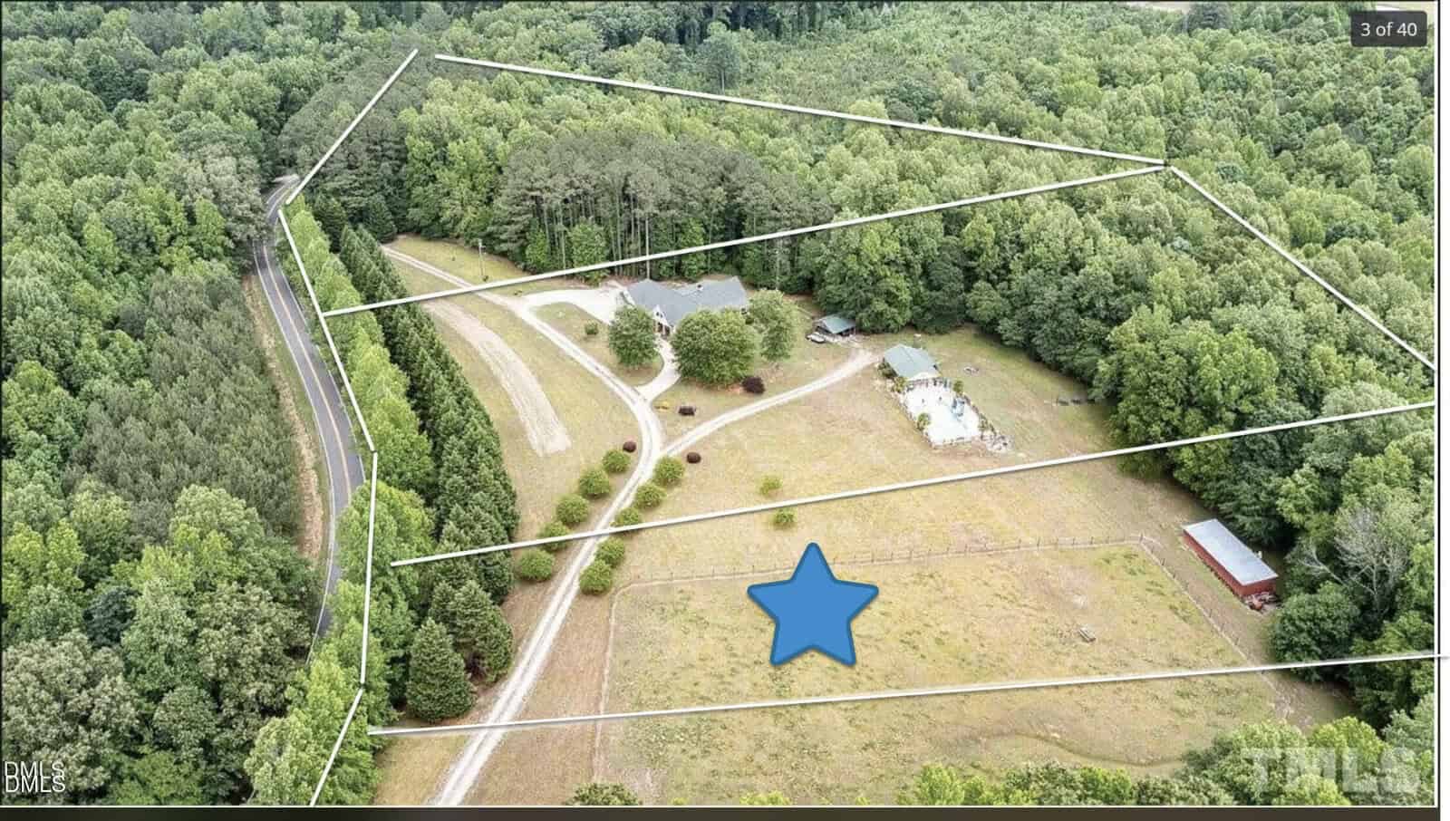 Aerial view of a rural property with wooded areas, open fields, a house, outbuildings, and a star marking a spot in a grassy area; property boundaries are outlined in white.