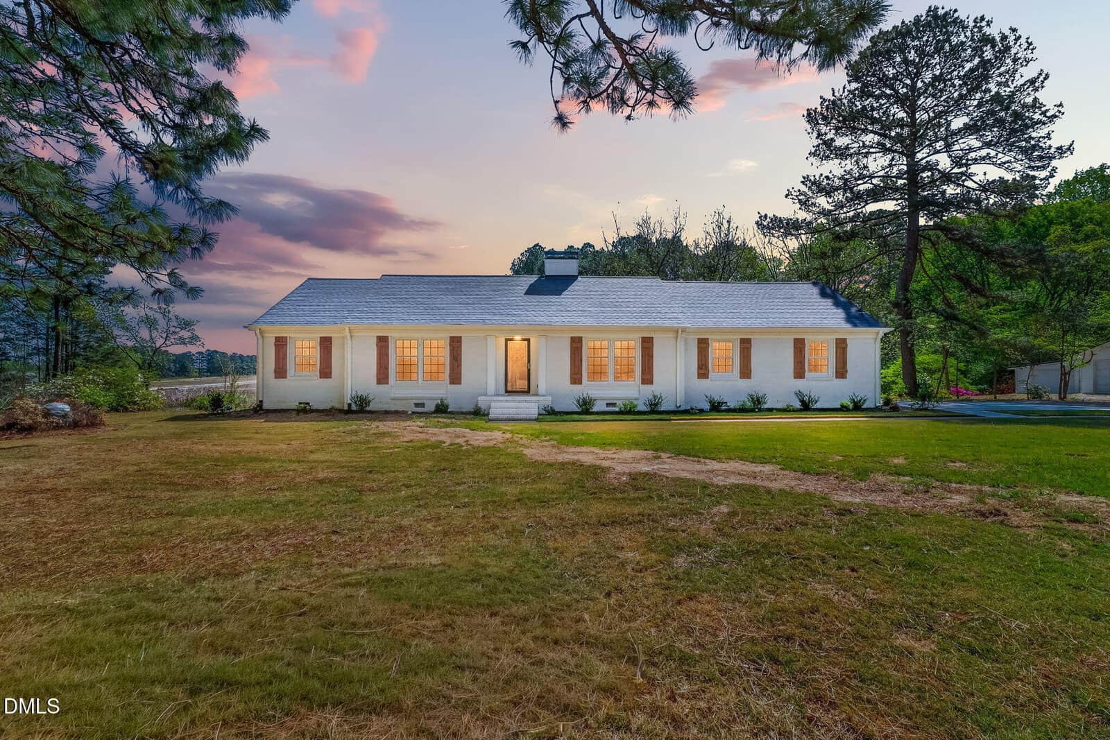 A single-story white house with a gray roof sits on a large grassy yard, surrounded by trees at sunset. Warm lights glow from the windows, creating a welcoming atmosphere under a colorful sky.