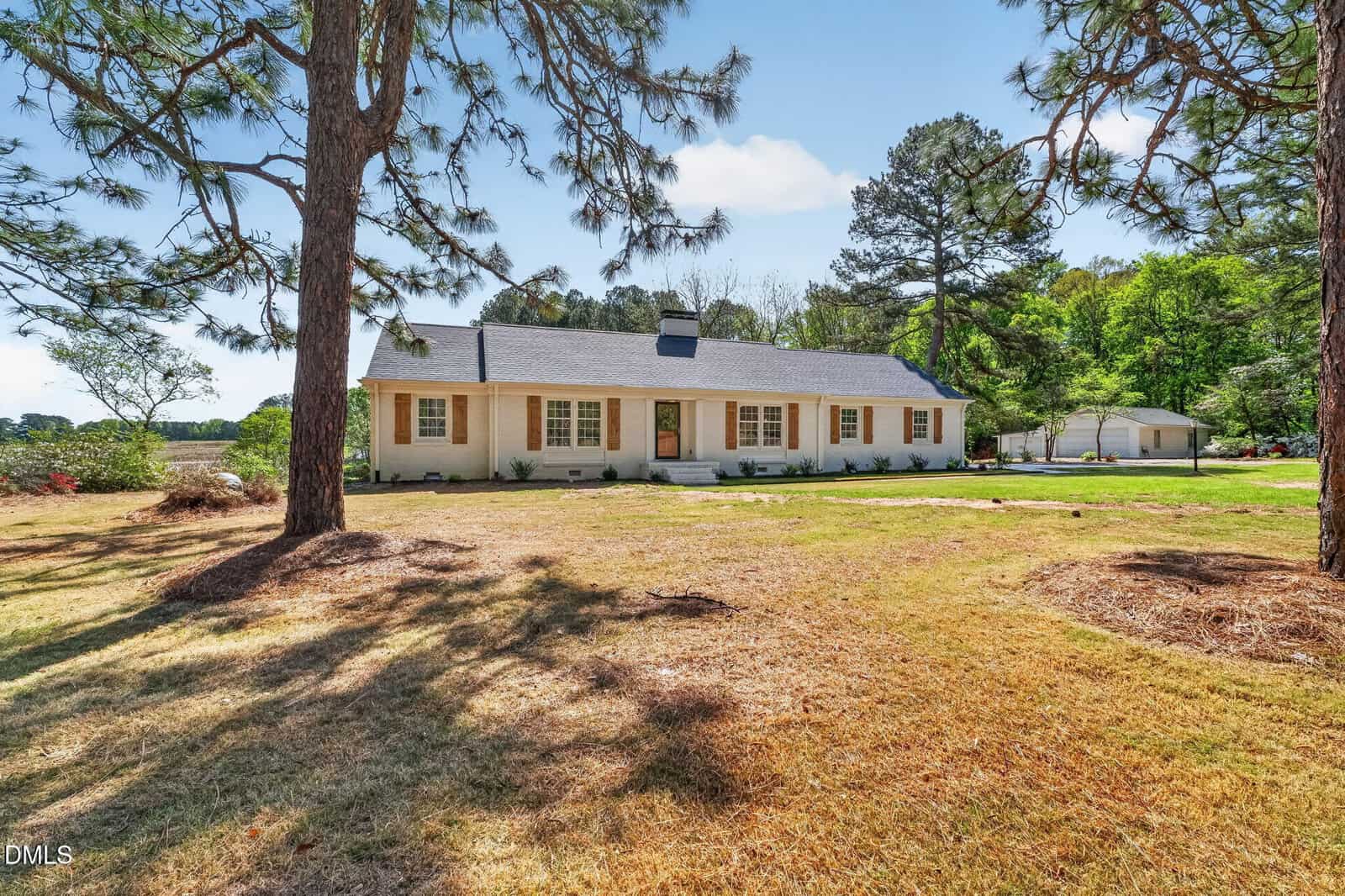 One-story white house with a dark roof, multiple windows with brown shutters, a front lawn with sparse grass, and large trees, set in a spacious, open yard with a detached white garage in the background.
