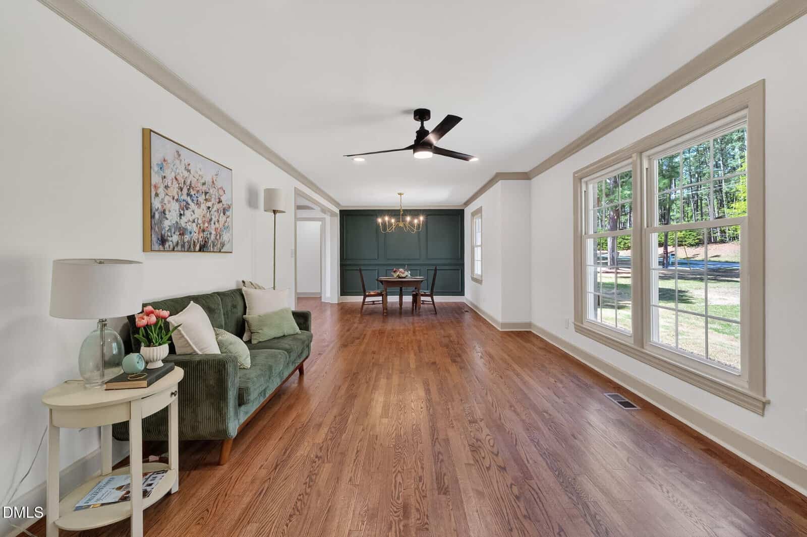 Spacious living and dining room with hardwood floors, large windows, a green sofa, side table, wall art, and a dining table under a modern chandelier, featuring white walls and beige trim.