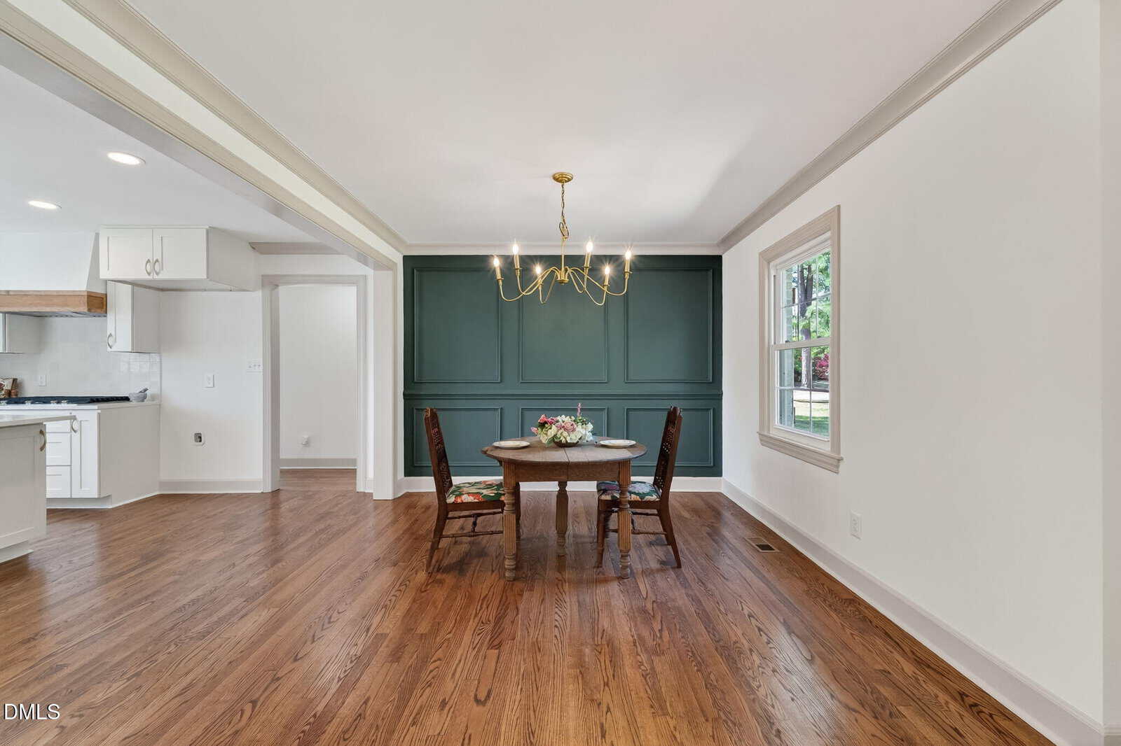 A dining area with a round wooden table, two chairs, a modern gold chandelier, dark green accent wall with molding, hardwood floors, and an adjacent kitchen with white cabinets. Natural light enters through a window.
