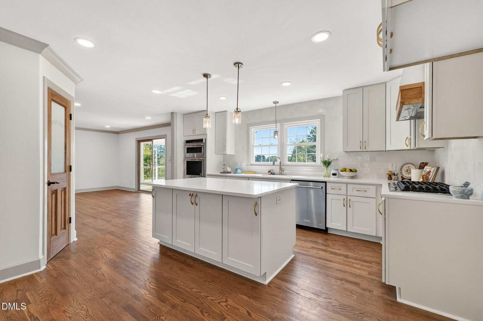 Bright, modern kitchen with white cabinets, a large island, stainless steel appliances, wood floors, pendant lighting, and a window over the sink. Sliding glass doors lead to a green outdoor area.