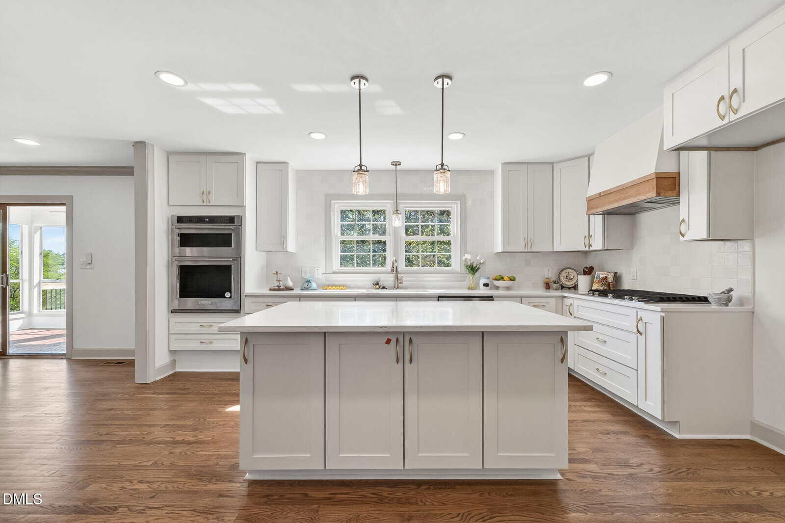 Bright, modern kitchen at 631 NC 39 Highway S, Louisburg features white cabinets, a large center island, stainless steel appliances, wooden floor, pendant lights, and a sunlit window. Glass doors open to an inviting outdoor area.