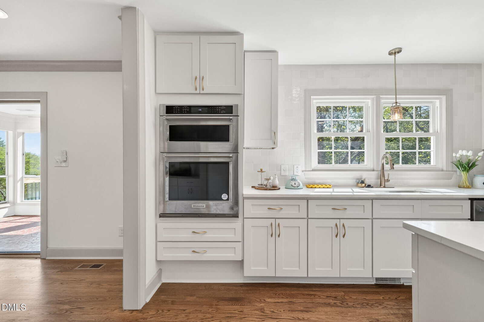 Bright kitchen with white cabinets, double wall oven, and large windows above a sink. Wooden floors and gold hardware add warmth. Sunlight fills the space, highlighting clean countertops and a vase of tulips by the window.
