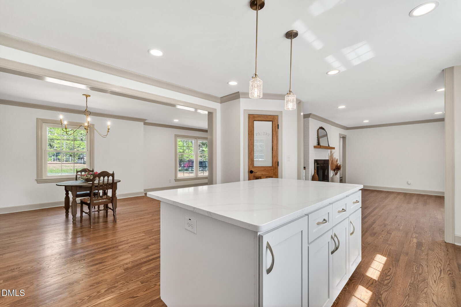Bright, open kitchen with white island countertop, pendant lights, hardwood floors, dining area with wooden table and chairs, and living room with fireplace. Large windows provide natural light.