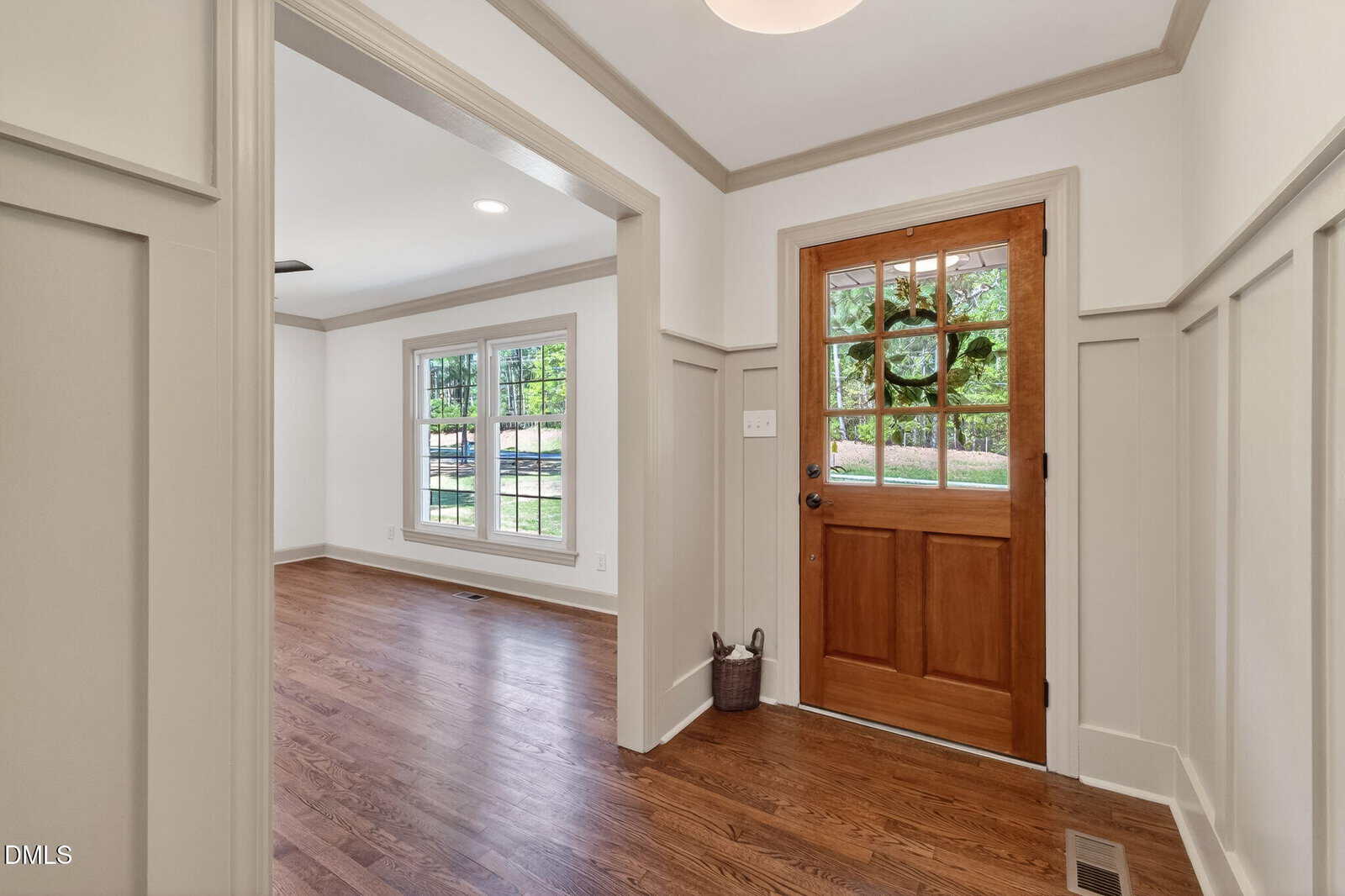 Bright entryway with natural wood front door, sidelights, wood flooring, white walls with wainscoting, and view into an adjoining room with large windows letting in natural light.