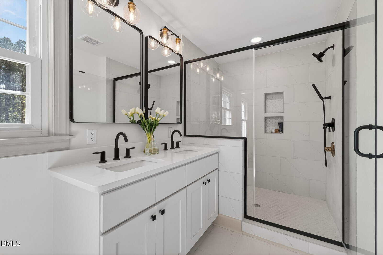 Modern bathroom at 631 NC 39 Highway S, Louisburg, featuring a double white vanity, black fixtures, large mirrors, a vase of white tulips, and a spacious glass-enclosed shower with white tile. Bright natural light fills the room.