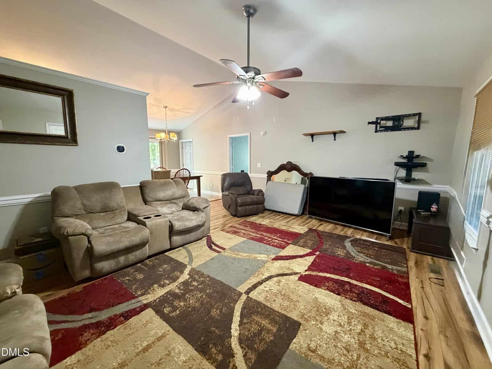 Spacious living room with beige recliners, patterned rug, ceiling fan, large TV on a stand, and hardwood floors. Dining area and kitchen are visible in the background.