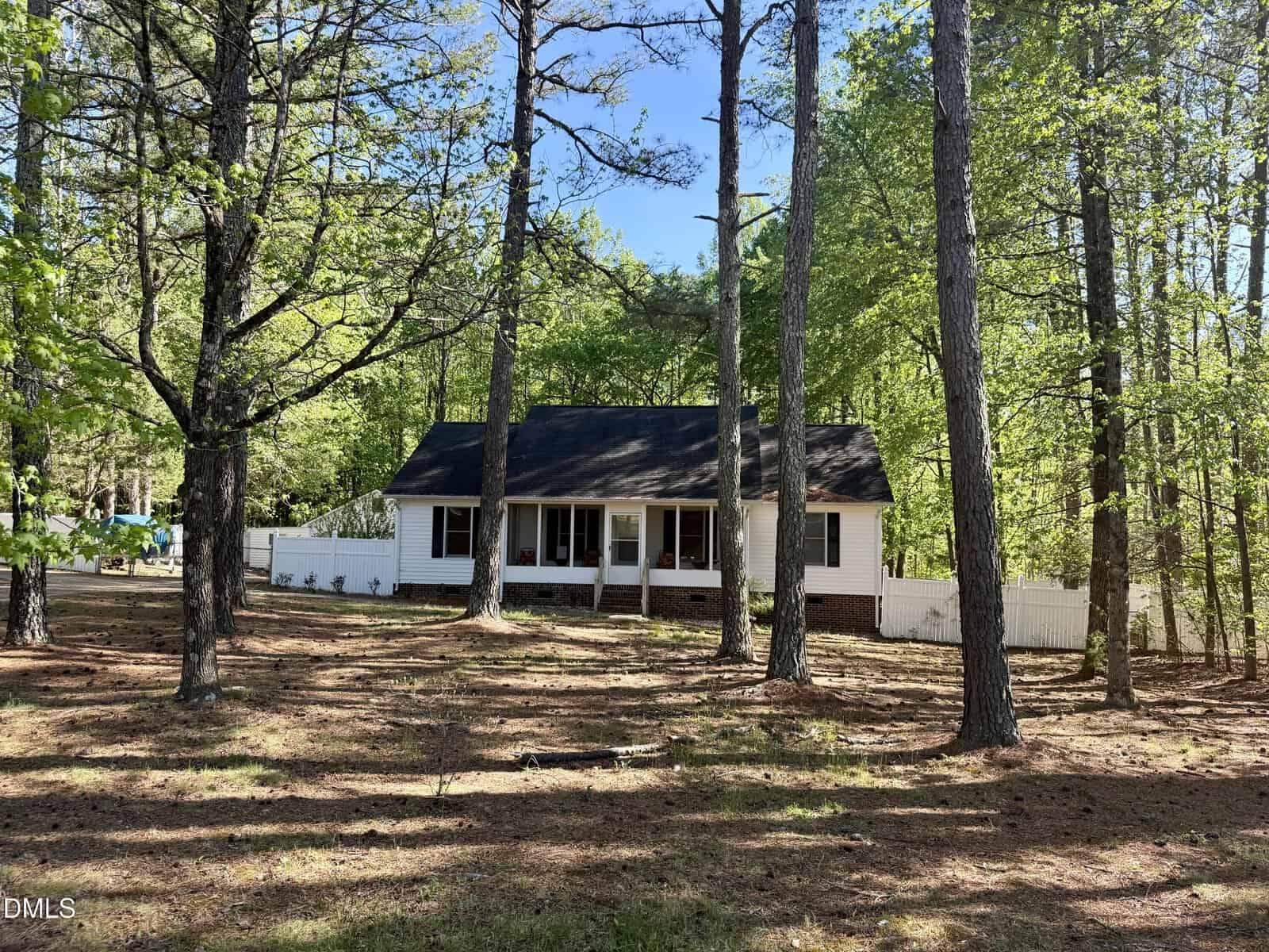 A small white house with a porch and dark roof sits among tall pine trees, surrounded by a white fence and green foliage under a clear blue sky.