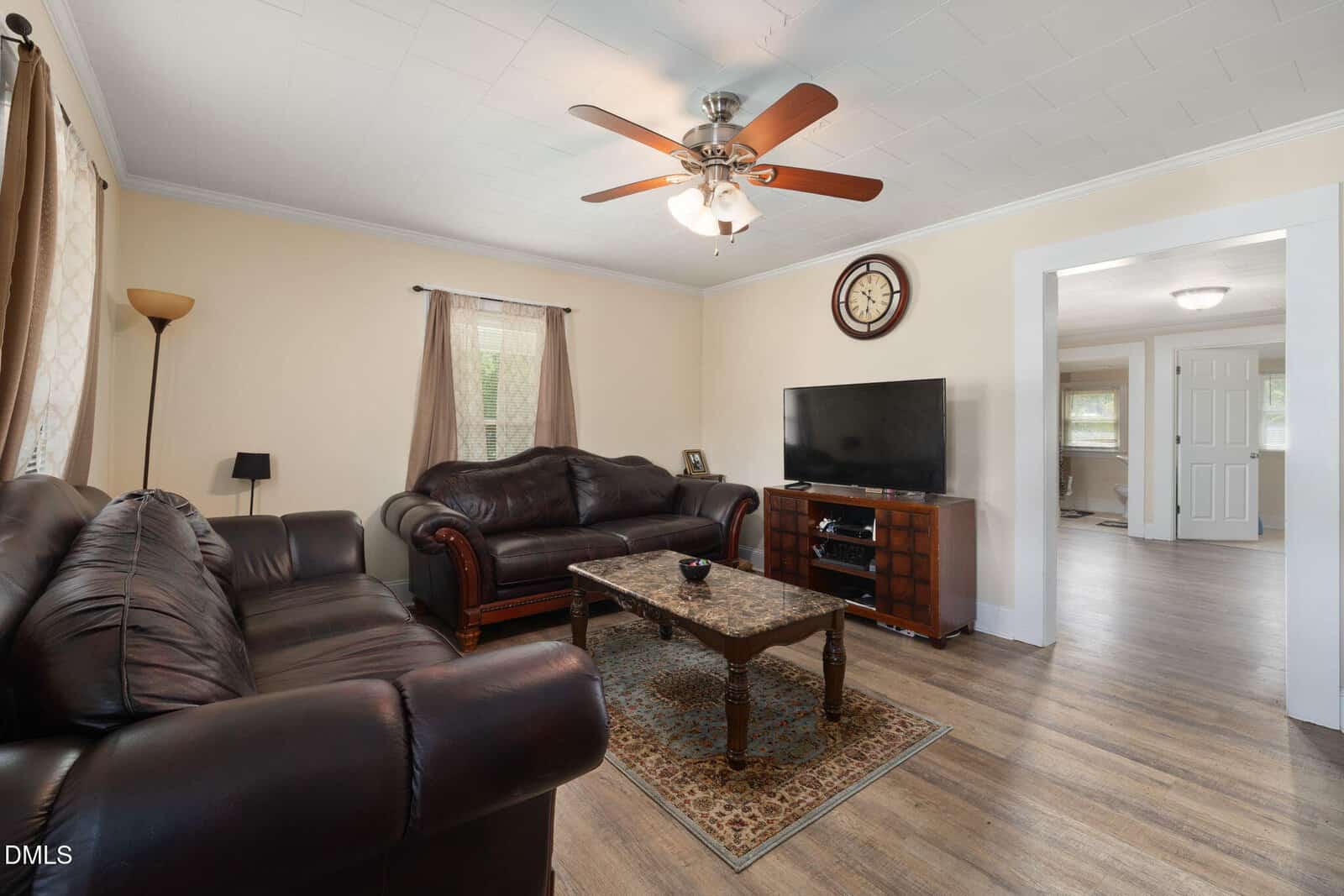 A living room with two dark leather sofas, a coffee table on a rug, a TV on a wooden stand, a ceiling fan, a wall clock, and light-colored walls and floors. Doorways lead to other rooms.