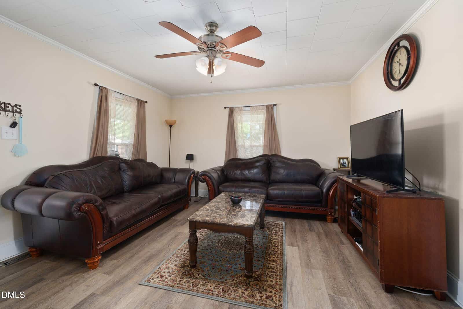 A living room with two dark leather sofas, a wooden coffee table, a rug, a TV on a stand, a clock on the wall, and light-colored walls and curtains. Ceiling fan with lights is centered above.