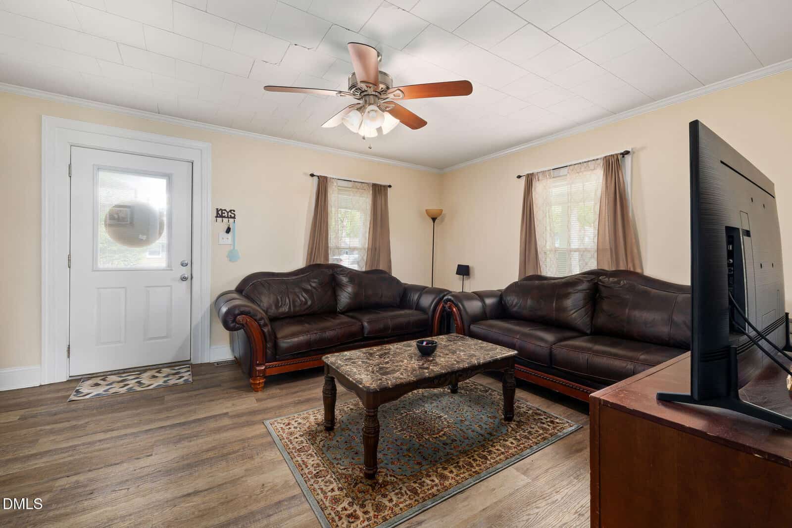 Living room with two dark leather sofas, a patterned rug, a wooden coffee table, a TV on a stand, a ceiling fan, and two windows with sheer curtains letting in natural light.