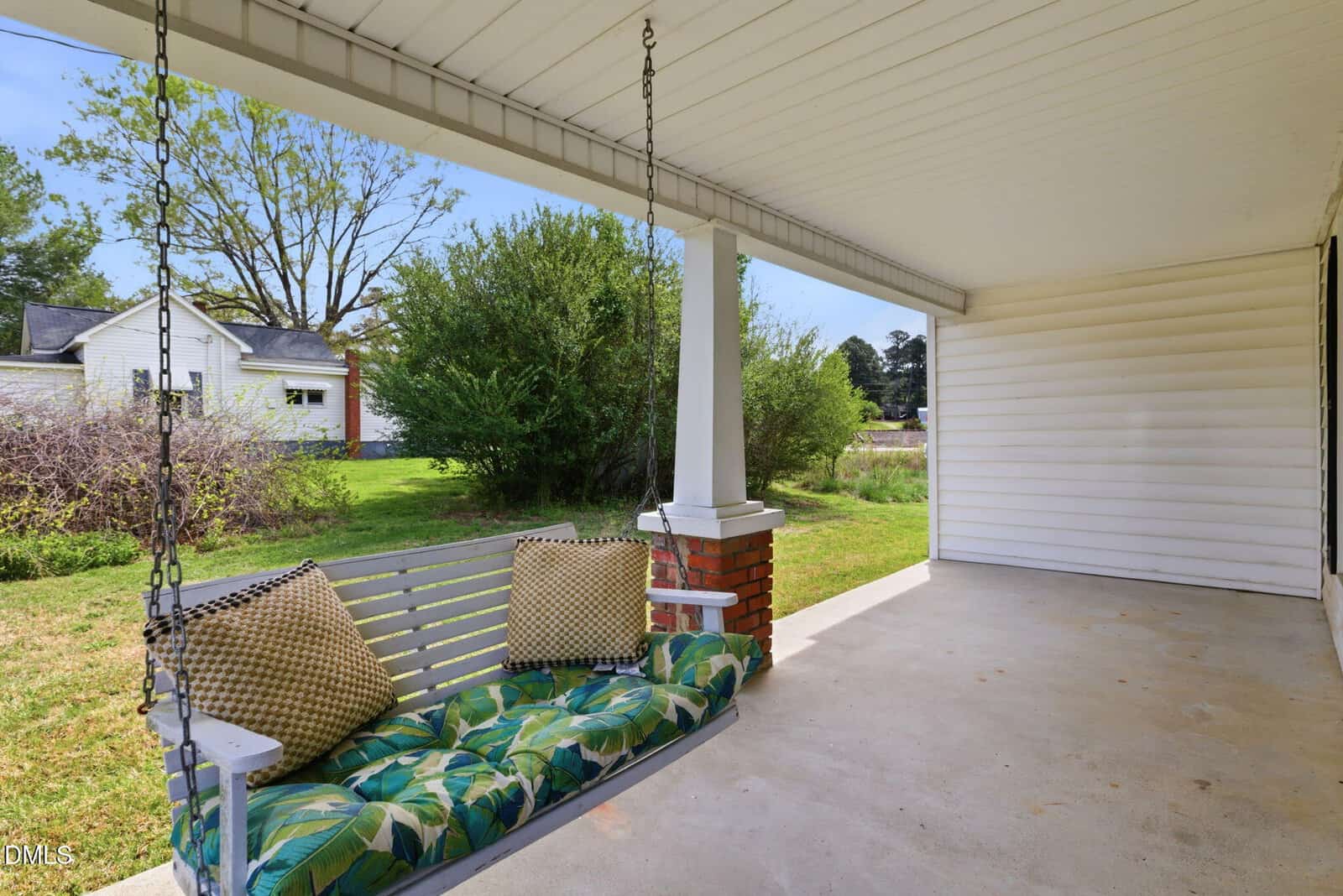 A covered front porch with a white porch swing adorned with green floral cushions and two pillows. The porch overlooks a grassy yard with trees and a white outbuilding in the background.