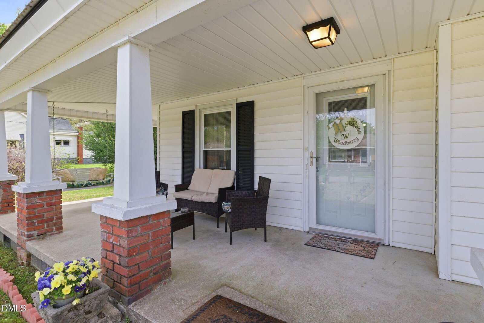 A cozy front porch with white siding, brick columns, two wicker chairs with cushions, a small glass table, and a decorative wreath on the glass door. Yellow flowers are planted along the edge.