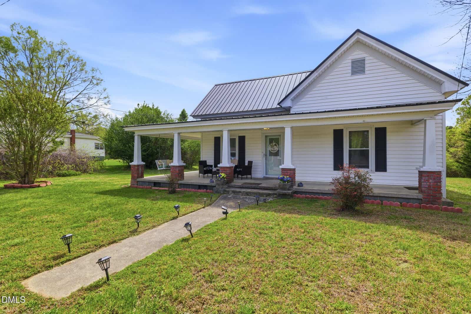 A white single-story house with a gray metal roof, covered front porch, brick pillars, and black shutters, surrounded by green grass, trees, and a clear blue sky. A concrete walkway leads to the porch.