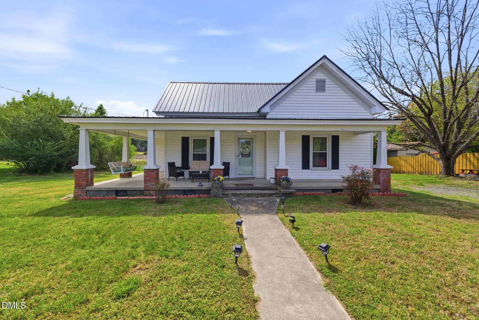 A white single-story house with a covered front porch, black shutters, and a metal roof. The porch has seating and a swing. A concrete walkway leads to the porch, and the lawn is bordered by bushes and trees.