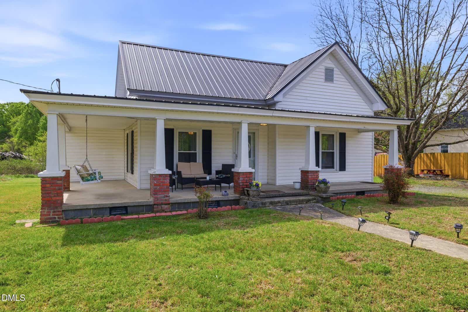 A white, single-story house with a metal roof, front porch, brick columns, outdoor furniture, and a swing, surrounded by green grass and trees, on a sunny day.