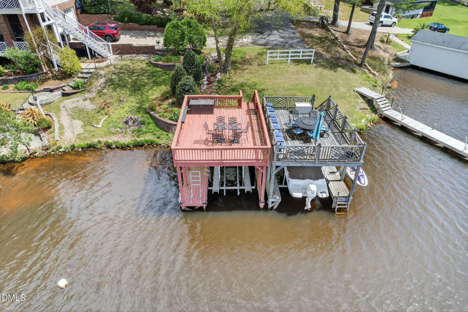 Aerial view of a lakeside dock with a covered patio and rooftop seating area, next to a deck with outdoor furniture and boats moored below, surrounded by grassy yard and nearby houses.