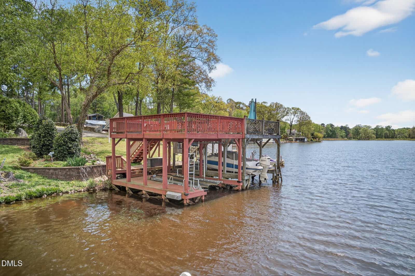 A wooden dock with a covered boat slip and an upper deck overlooks a calm lake, surrounded by green trees and grassy banks under a partly cloudy blue sky.
