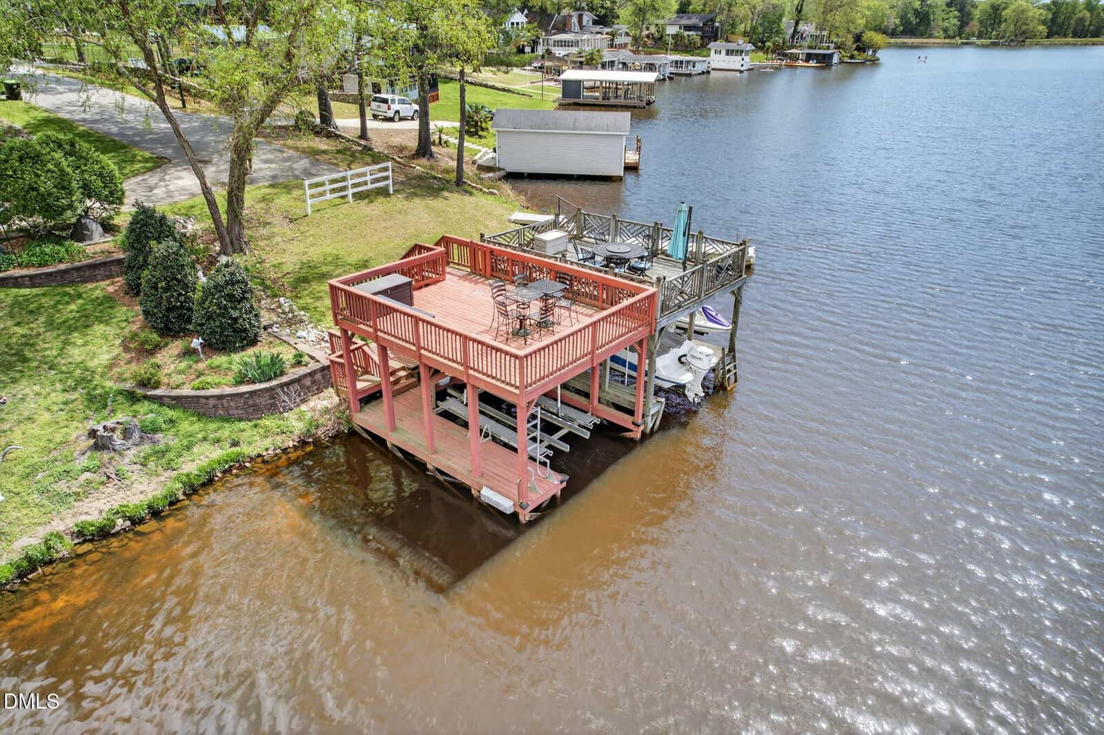 A two-level red wooden dock with outdoor seating overlooks a calm lake at 30 Ray Avenue, Zebulon, surrounded by green lawns, trees, and waterfront houses on a sunny day.