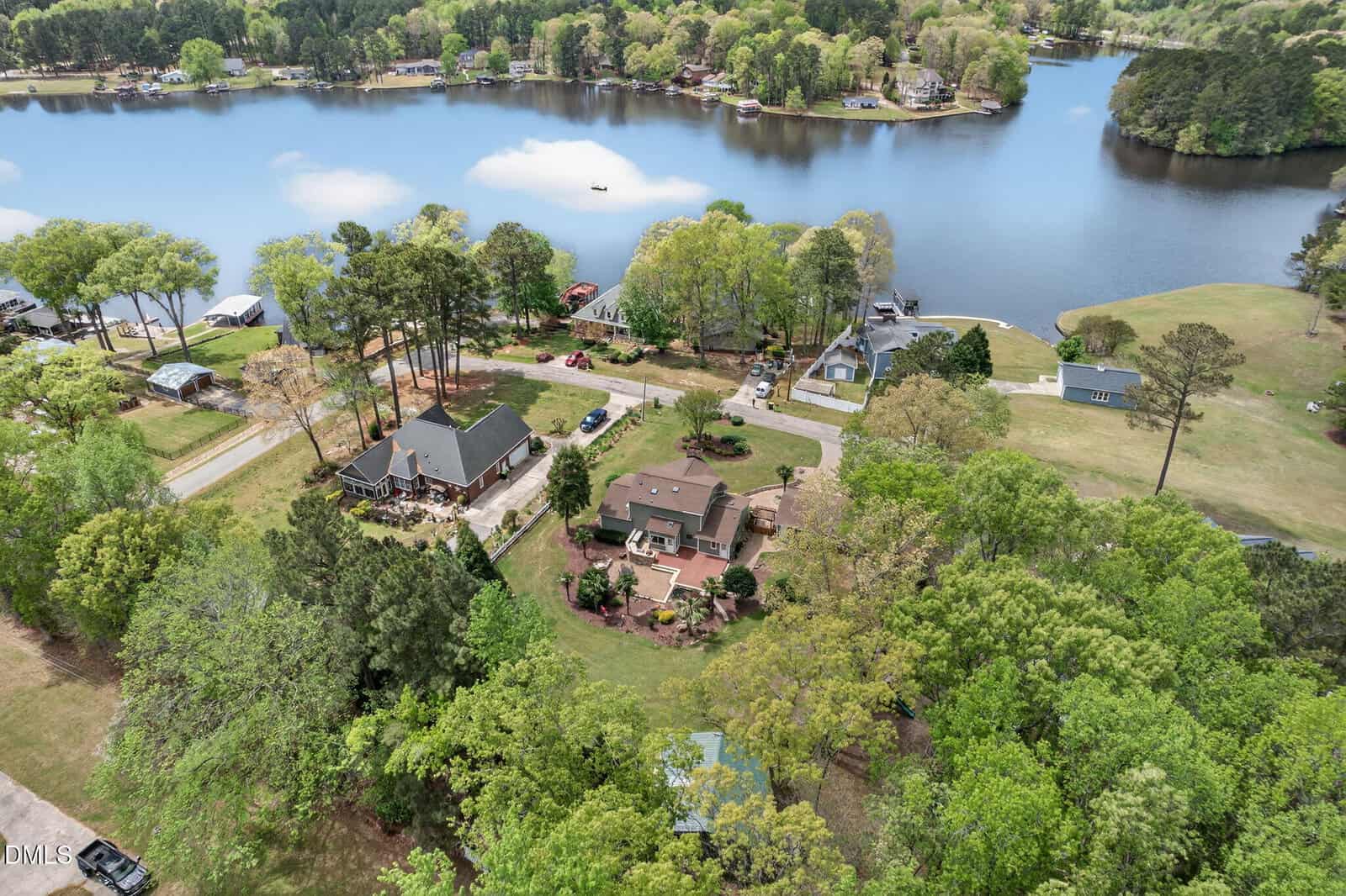 Aerial view of a lakeside neighborhood with several houses surrounded by trees, well-kept lawns, and docks along the calm water, under a partly cloudy sky.
