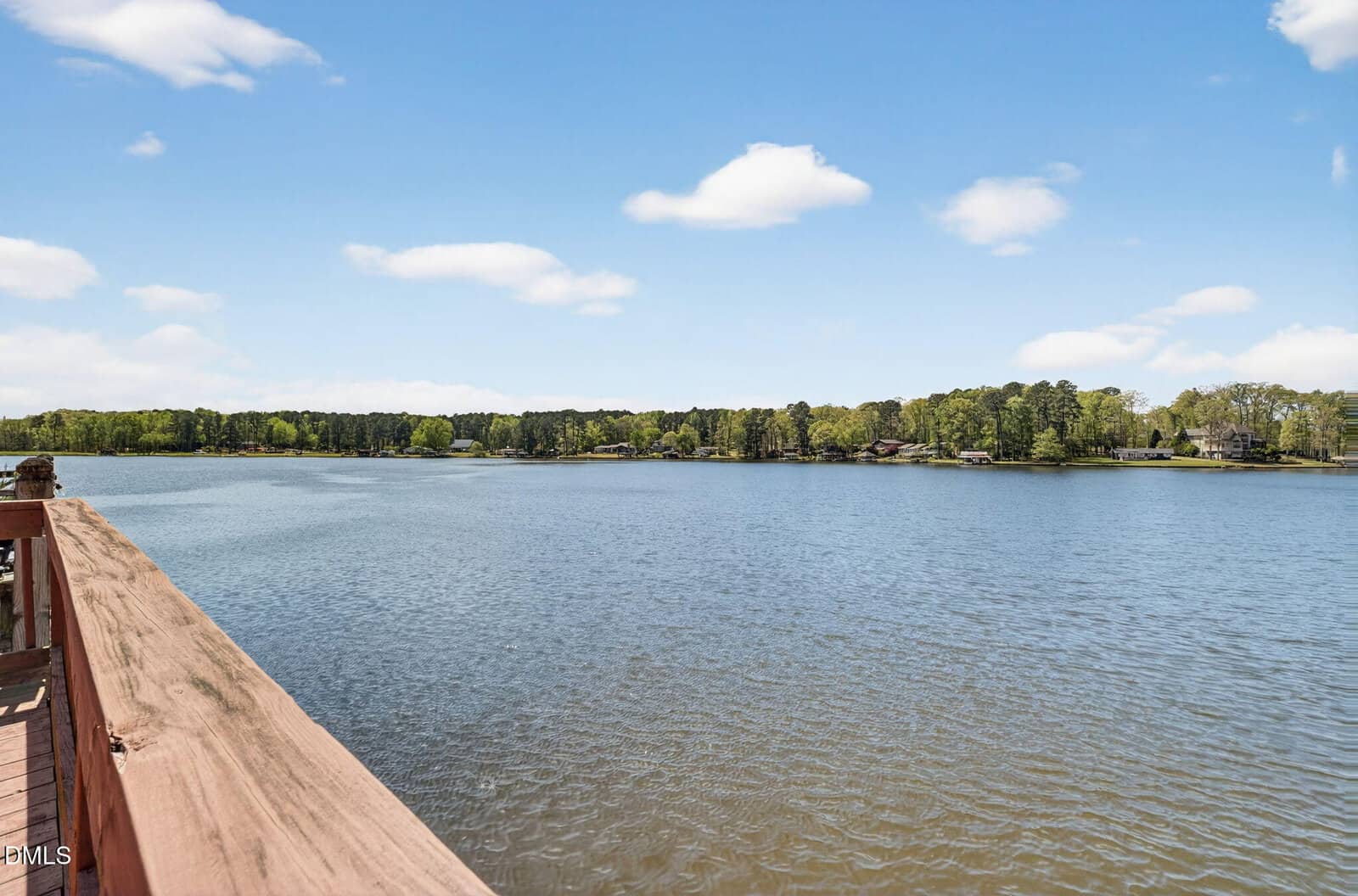 View from a wooden deck overlooking a wide, calm lake with a tree-lined shore under a clear blue sky with a few scattered clouds.