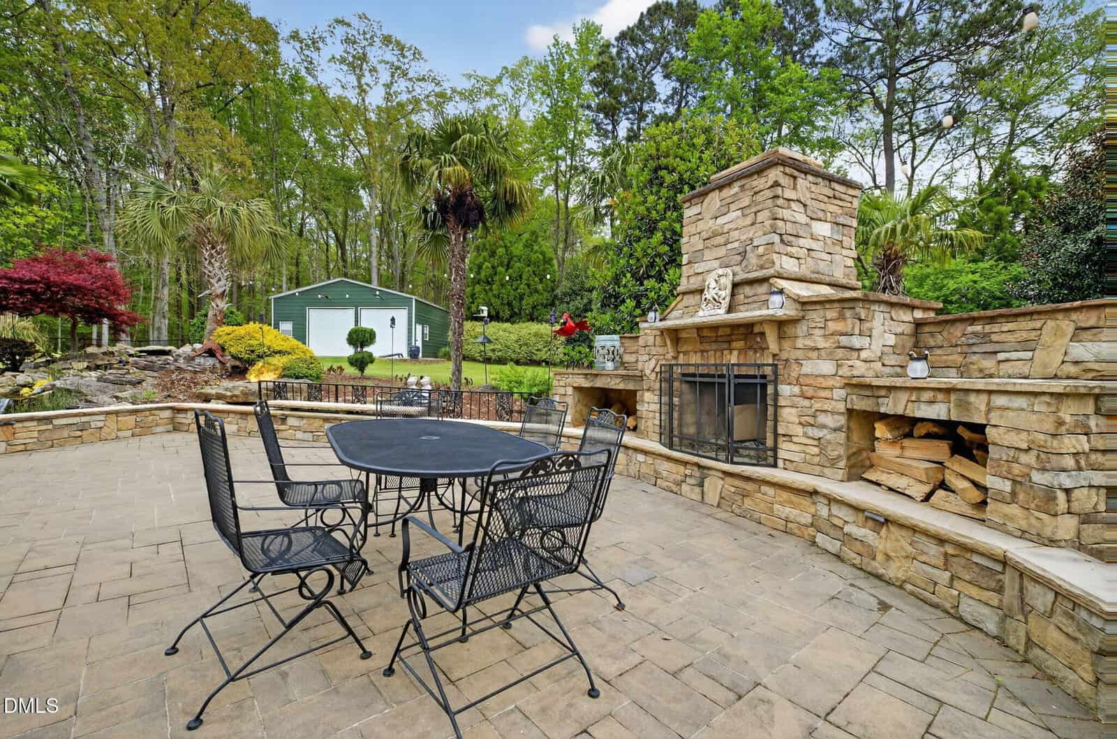 A stone patio with a black metal table and chairs, a large stone outdoor fireplace with stacked firewood, surrounded by lush greenery, palm trees, and a green shed in the background.