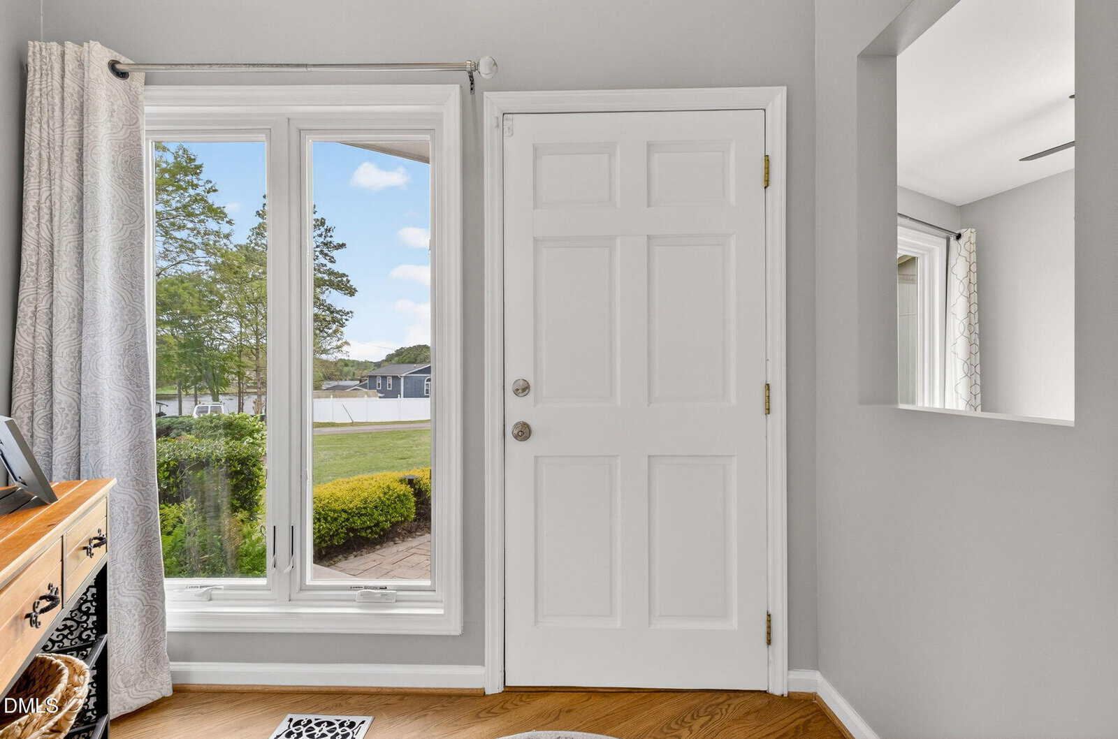A white front door with a brass handle, next to large windows with gray curtains, overlooks a green yard and distant houses. A wooden desk and a mirror are visible inside.
