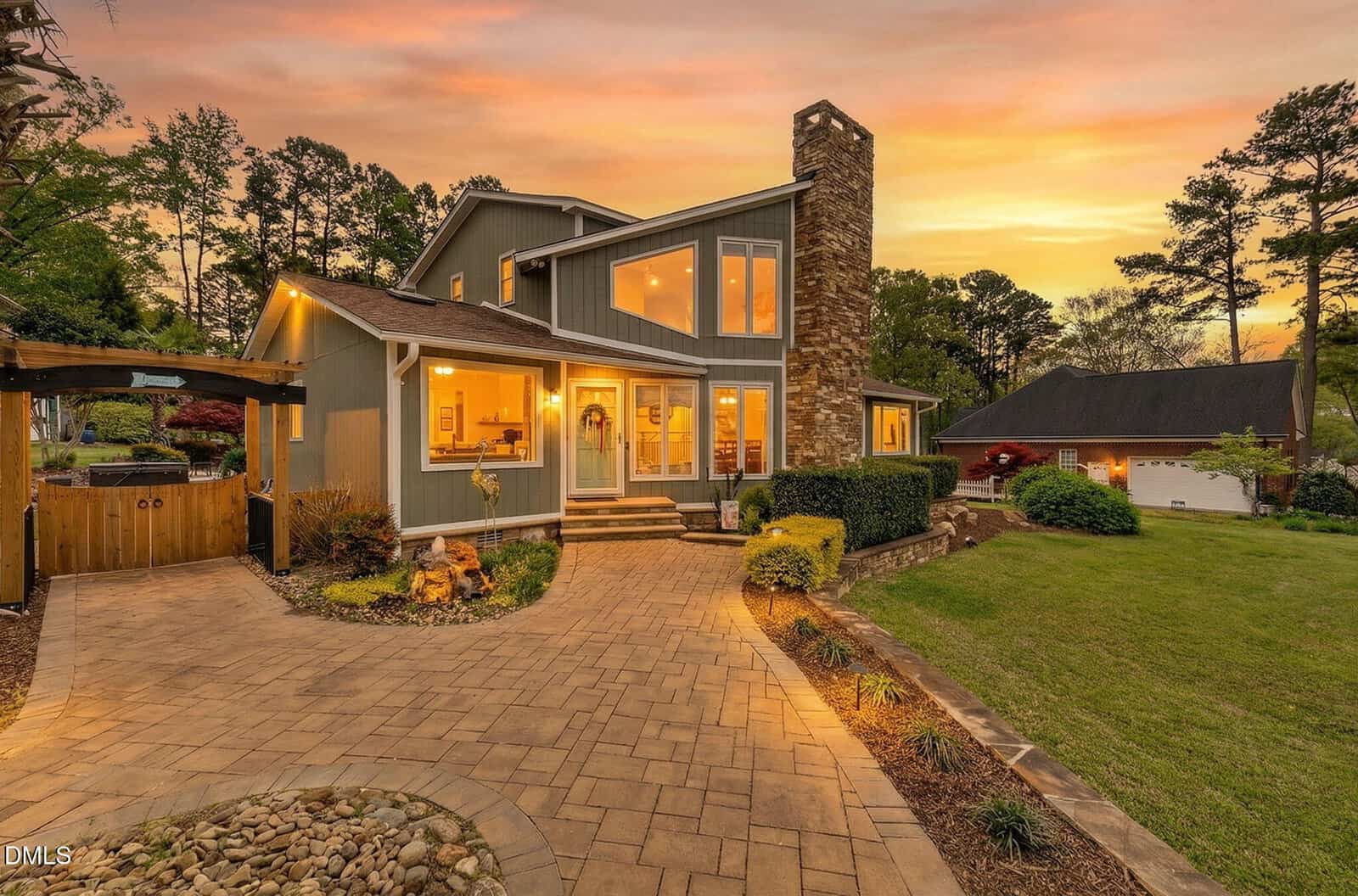 Modern house with large windows, stone chimney, and front porch at sunset. Warm exterior lights, a landscaped yard, and paved walkway enhance the cozy, inviting atmosphere. Trees and a detached garage are in the background.