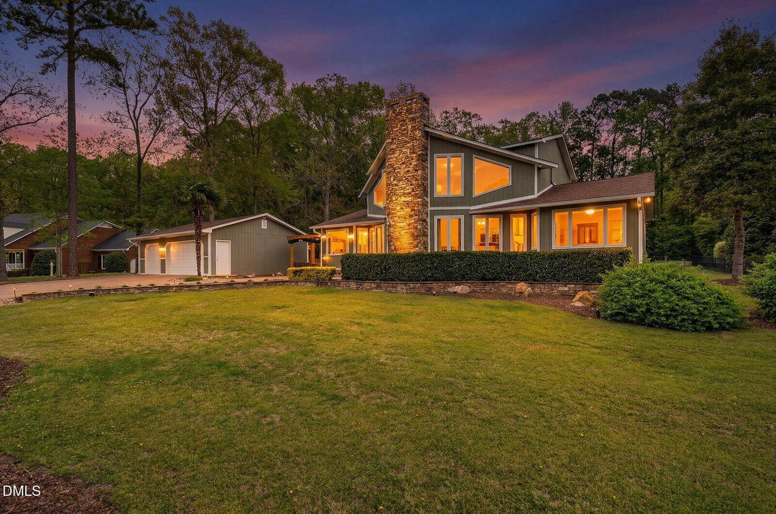 A modern two-story house with large windows and a stone chimney, warmly lit at dusk. The home features a spacious front lawn, a detached garage, and is surrounded by tall trees and landscaped shrubs.