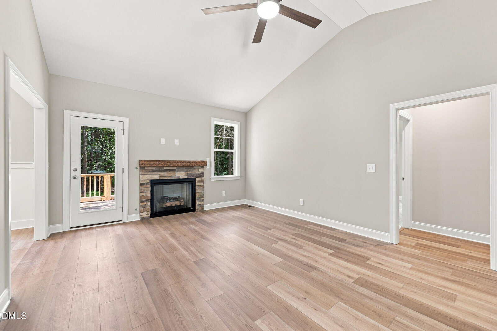 A spacious living room with light wood flooring, a ceiling fan, a fireplace with a stone accent, a door and window leading outside, and entryways to other rooms. Walls are painted light gray.