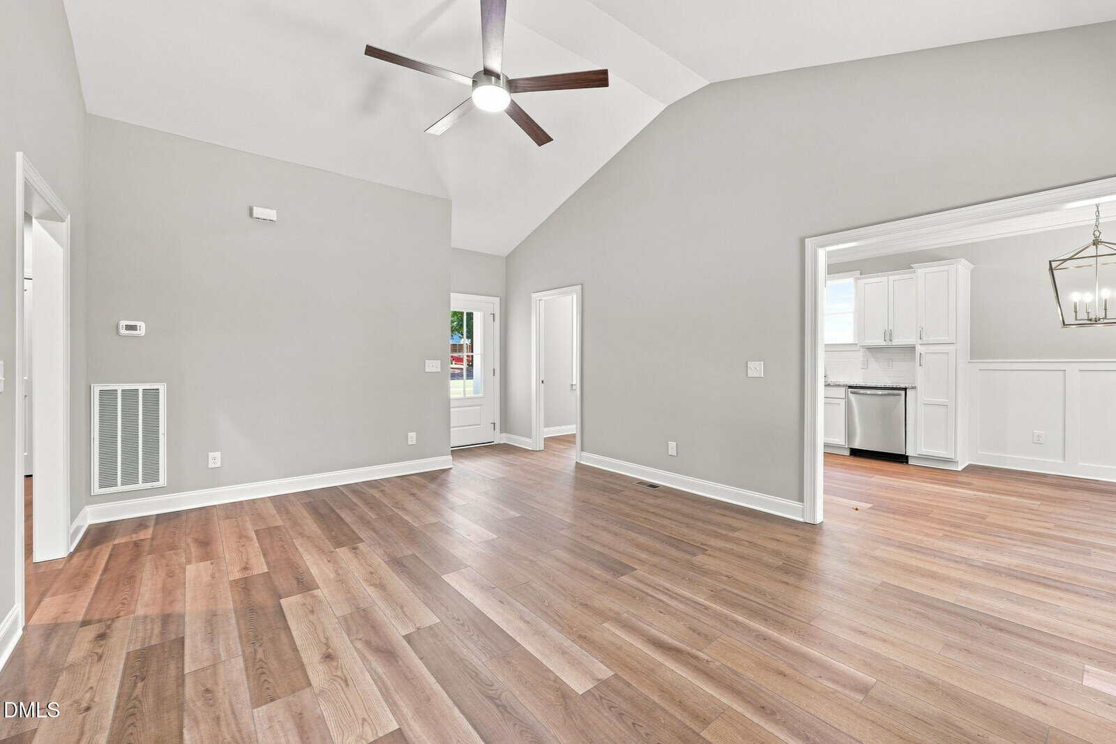 A spacious room with light gray walls, wood flooring, a ceiling fan, and vaulted ceiling. There are two doorways: one leading to a kitchen with white cabinets, and another to a hallway or room.