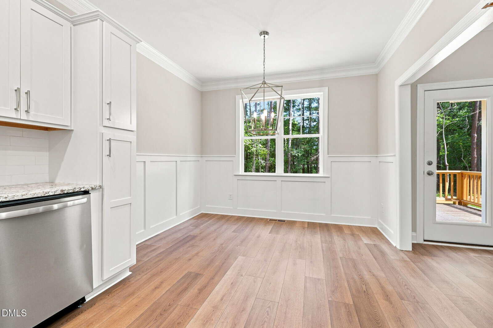 Bright kitchen area with white cabinets, light wood flooring, crown molding, a window overlooking trees, a geometric light fixture, and a glass door leading to an outdoor deck.
