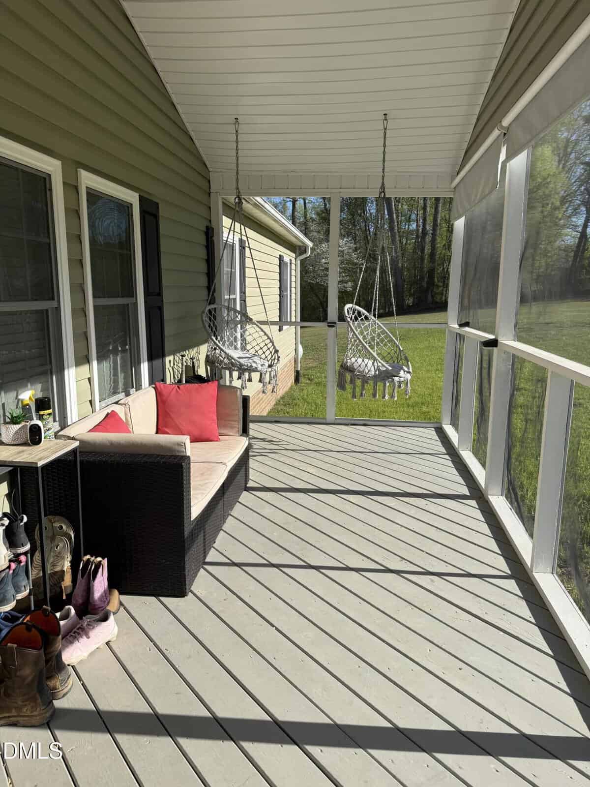 A screened-in porch with a wicker loveseat, red cushions, two hanging basket chairs, a boot rack with shoes, and sunlight streaming across the wooden floor. Green lawn and trees are visible outside.