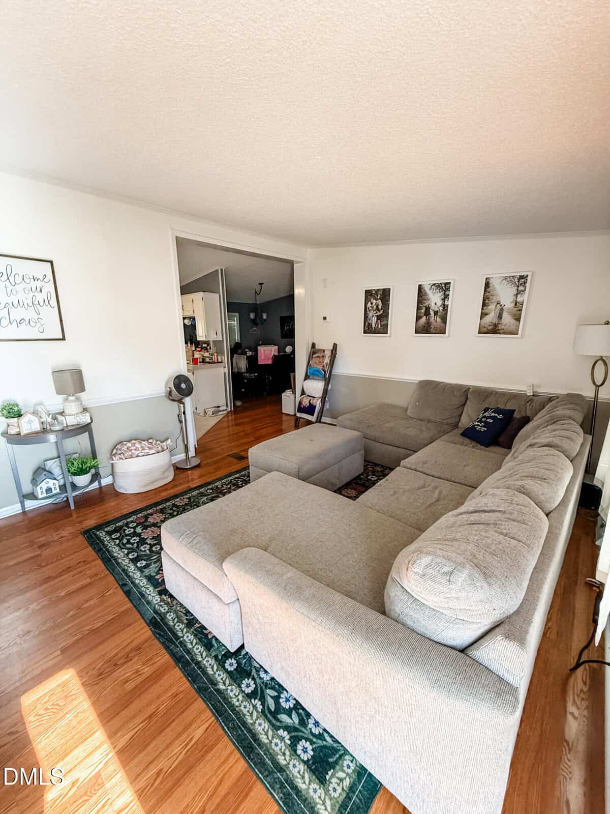 A cozy living room with a large beige sectional sofa, green patterned rug, wooden floors, and wall art. Decorative accents include a small round side table, fan, and houseplants. An entryway is visible in the background.
