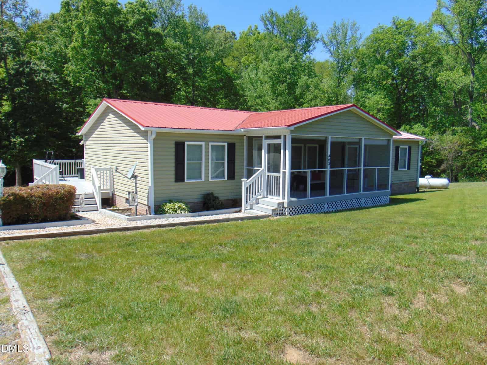 Single-story beige house with a red metal roof, screened front porch, ramp, and steps, surrounded by green grass and trees in the background under a clear blue sky.