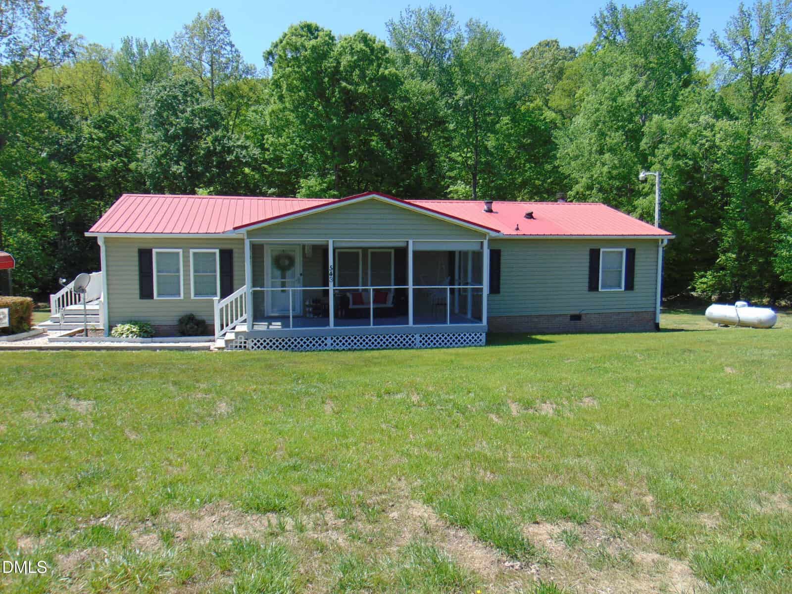Single-story house with light siding and a red metal roof, featuring a screened front porch and white steps, set on a large grassy yard with trees in the background on a sunny day.