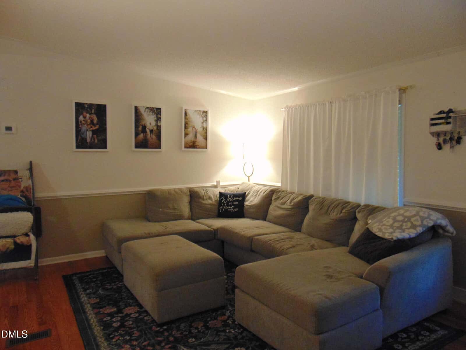 A cozy living room with a large L-shaped beige sectional sofa, three ottomans, a lamp, wall art, and a dark patterned rug. White curtains cover the window, and pillows decorate the sofa.