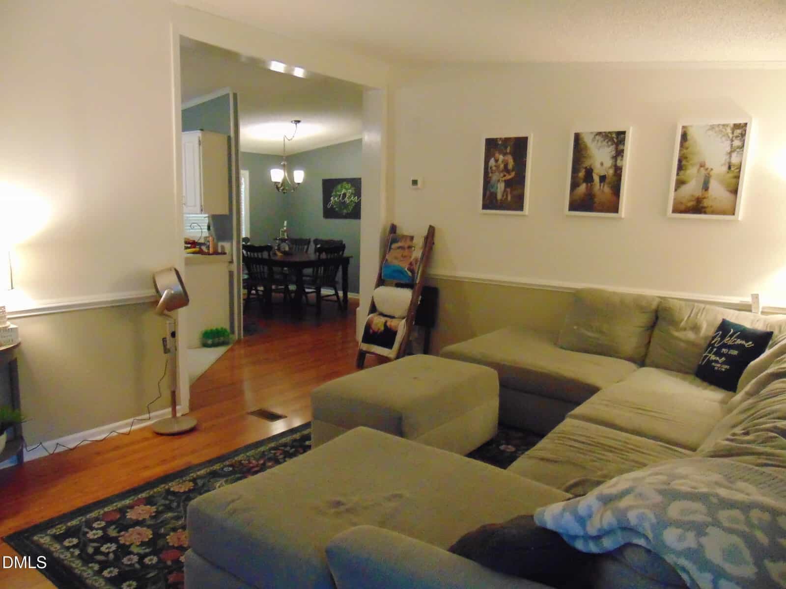 A cozy living room with a sectional sofa, ottoman, rug, and wall art. The room opens to a dining area with a wooden floor, table, and chairs. A floor lamp and fan are visible near the sofa.