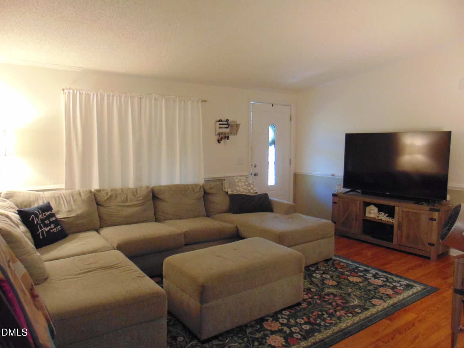 A cozy living room with a large gray sectional sofa, matching ottoman, a flat-screen TV on a stand, white curtains, a patterned rug, hardwood floors, and a door leading outside.