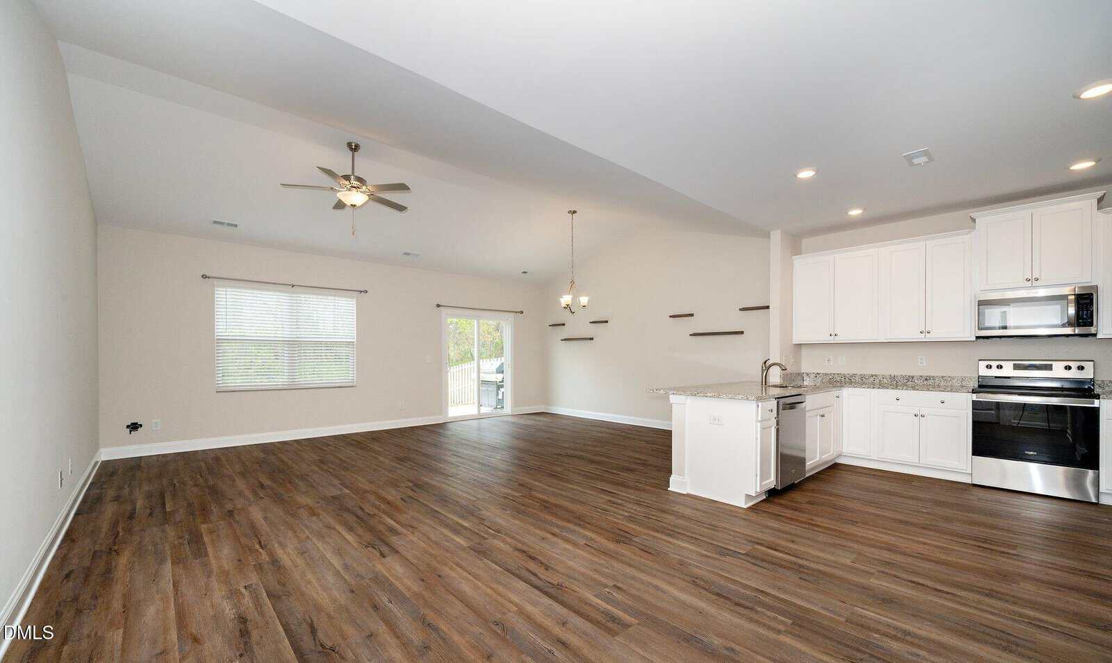 Open-concept living area with wood floors, white kitchen cabinets, stainless steel appliances, ceiling fan, large window, sliding glass door, granite counters, and floating shelves on the wall.