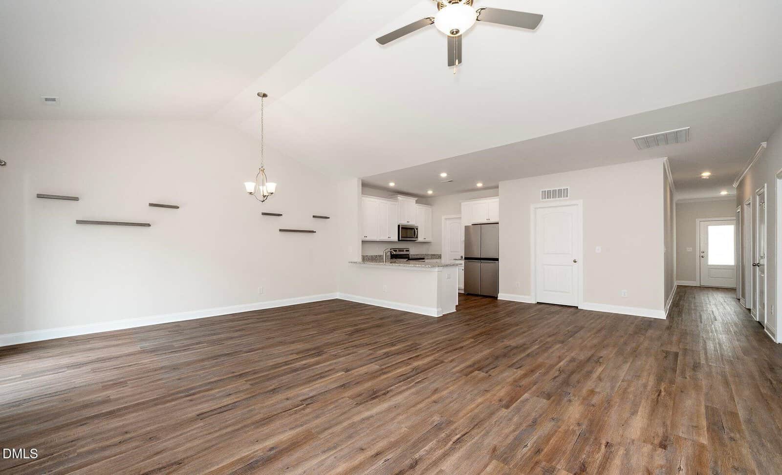 Empty, modern open-plan living space with wood flooring, white walls, a ceiling fan, kitchen area with stainless steel appliances, hanging lights, and built-in wall shelves. Hallway and doorways are visible in the background.