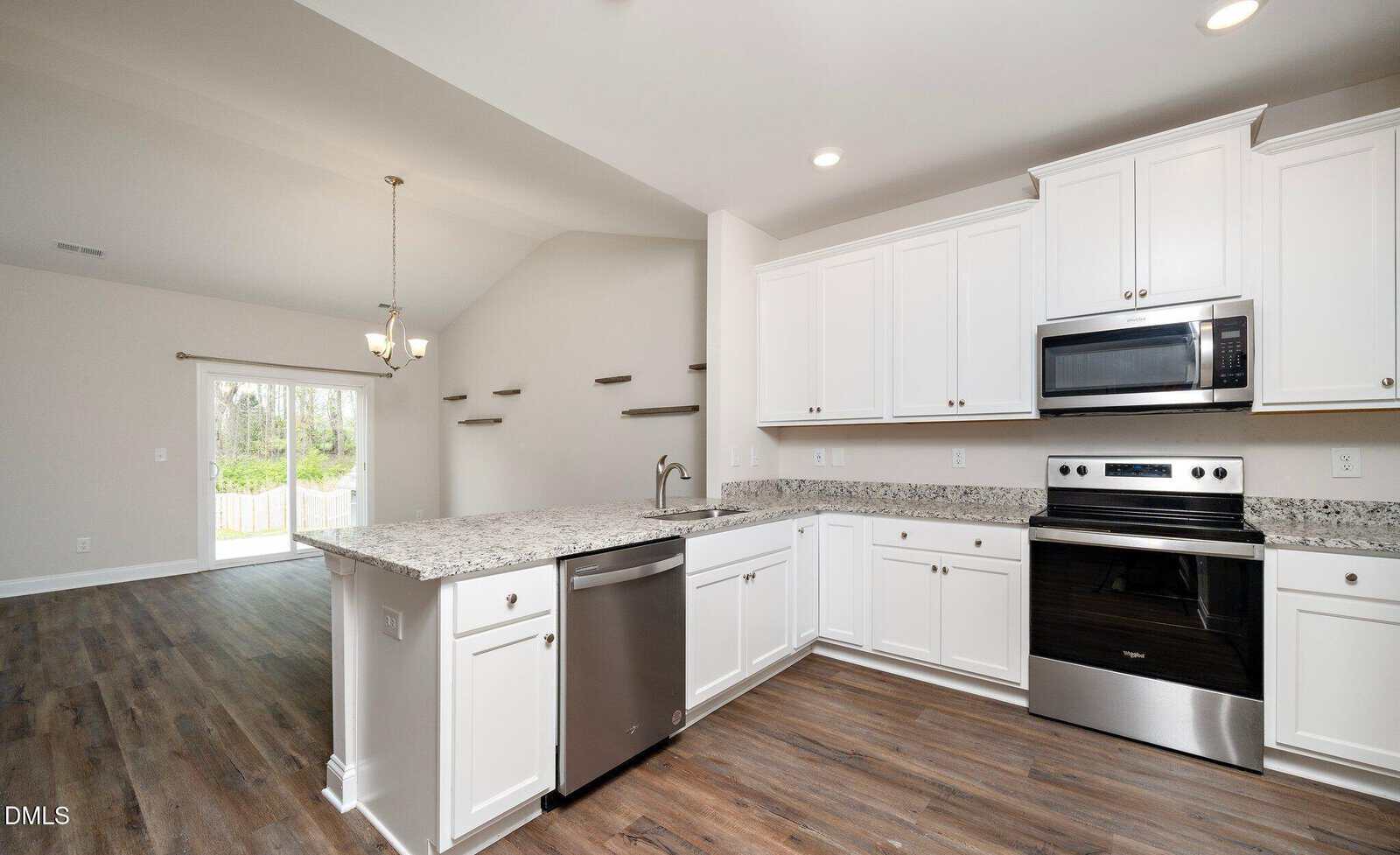 Modern kitchen with white cabinets, stainless steel appliances, granite countertops, and wood flooring; open layout leads to a dining area with shelves on the wall and glass doors opening to a backyard.