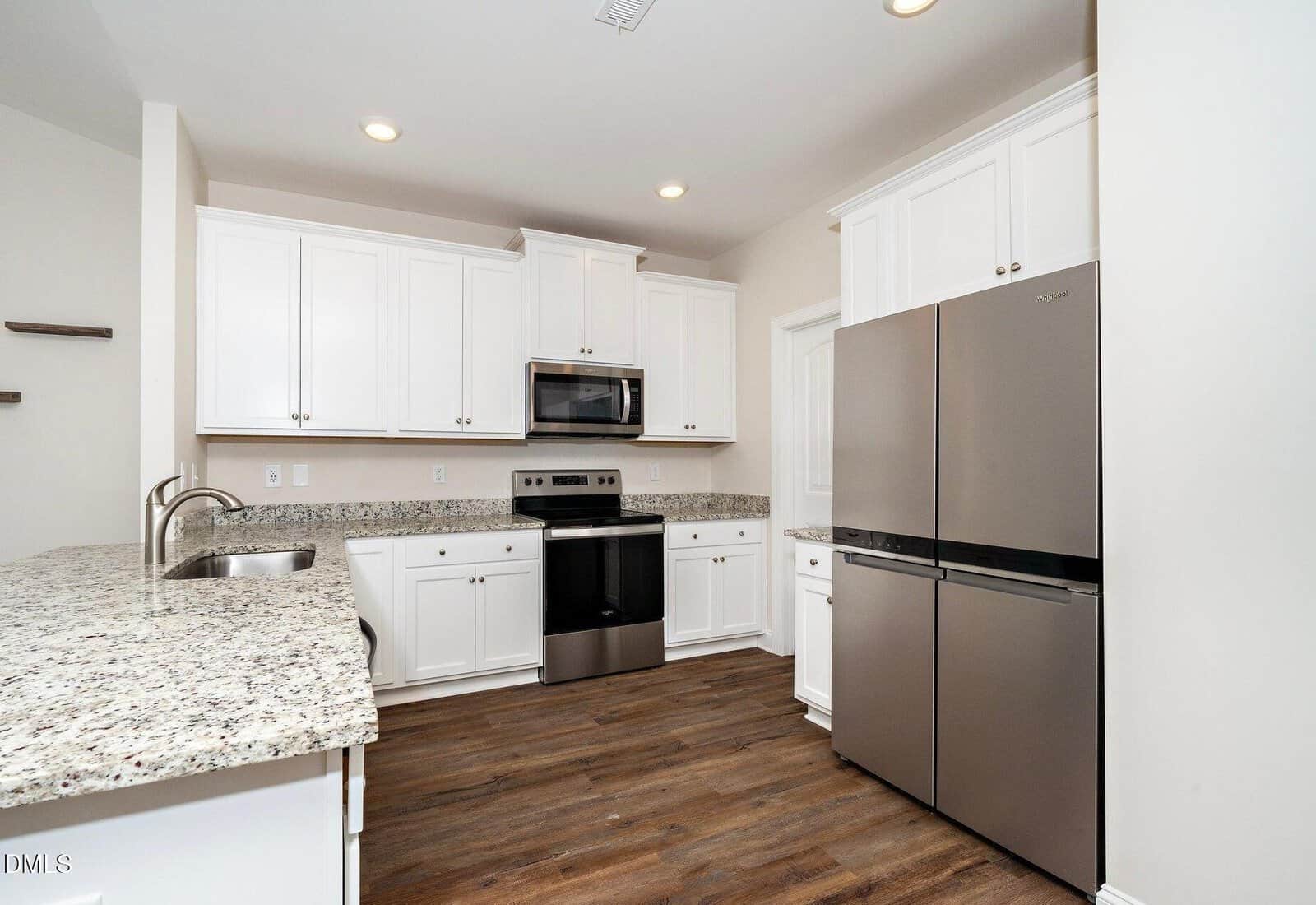 Modern kitchen with white cabinets, granite countertops, stainless steel appliances including a double-door refrigerator and oven, and wood-look flooring. The walls are light-colored, creating a bright, clean atmosphere.
