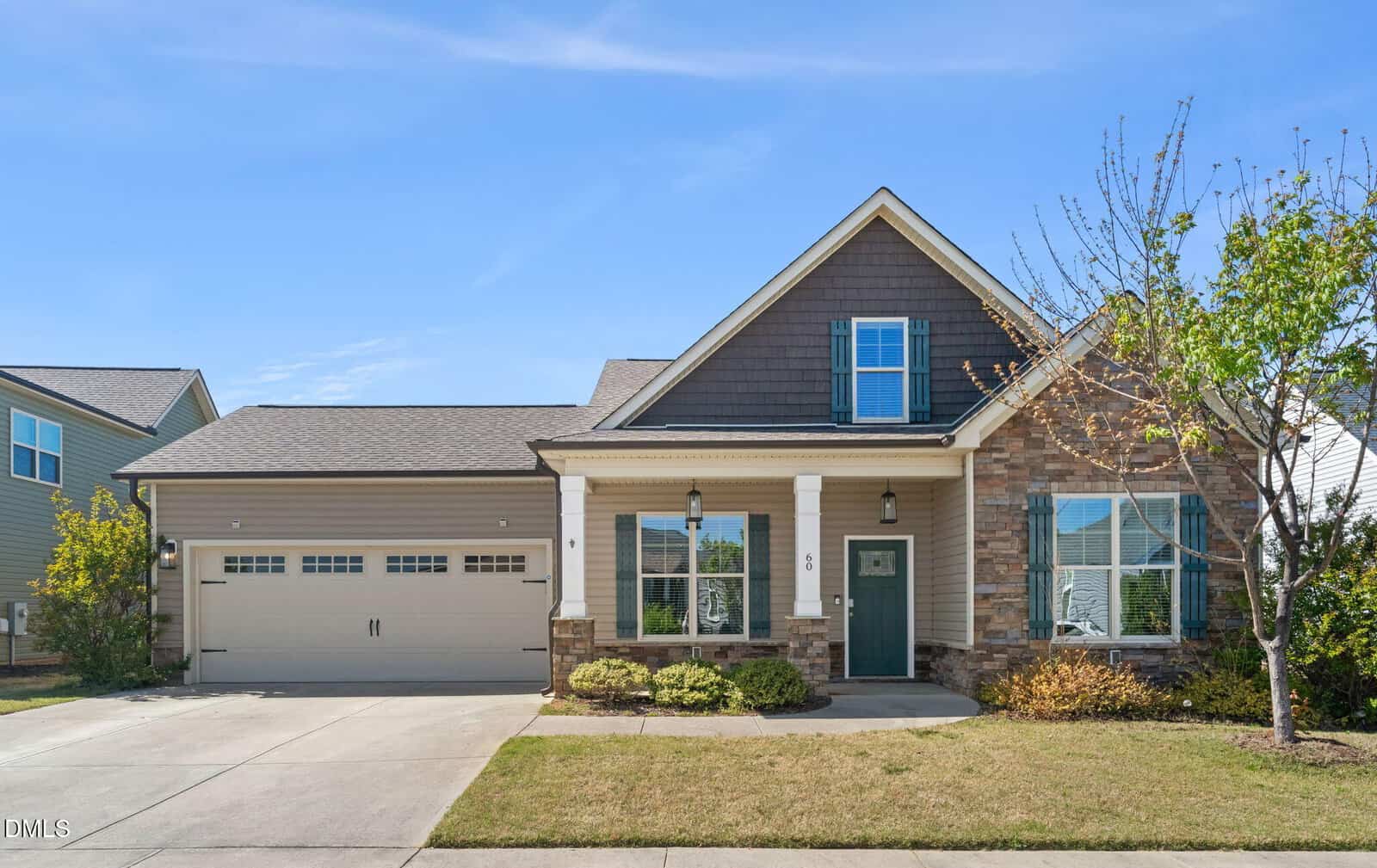 Single-story suburban house at 60 Scarlet Bell Drive in Youngsville with a two-car garage, stone and siding exterior, front porch with columns, green door, shuttered windows, and a small front yard with bushes and a tree, under a clear blue sky.